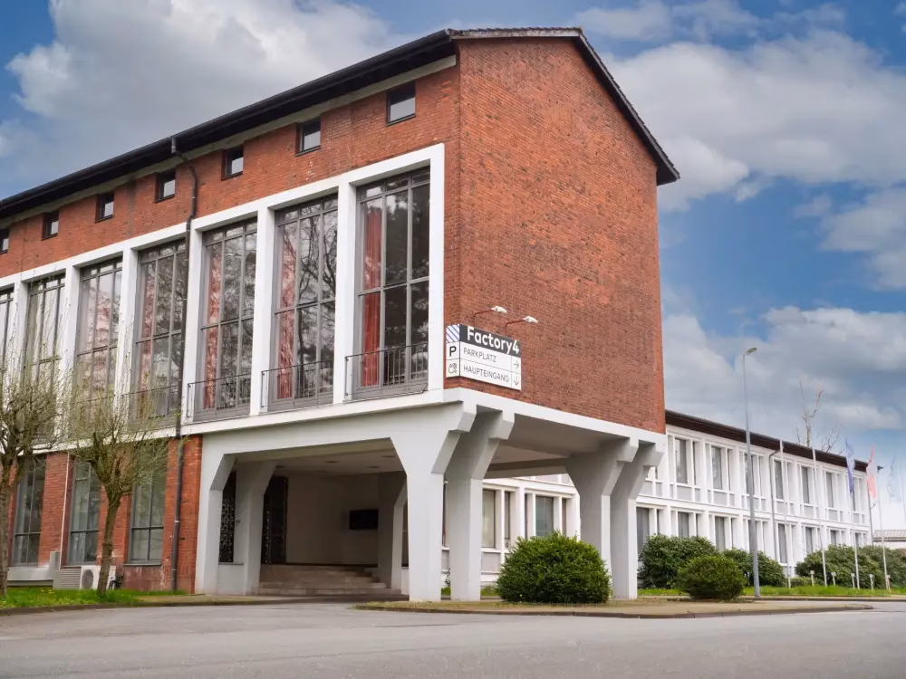 Das Firmengebäude von GrabGoose: ein modernes Backsteingebäude mit großen, weißen Fensterrahmen. An der Ecke ist ein Schild mit der Aufschrift „Factory4“ sowie Hinweisen zum Parkplatz und Haupteingang angebracht. Der Himmel ist leicht bewölkt.