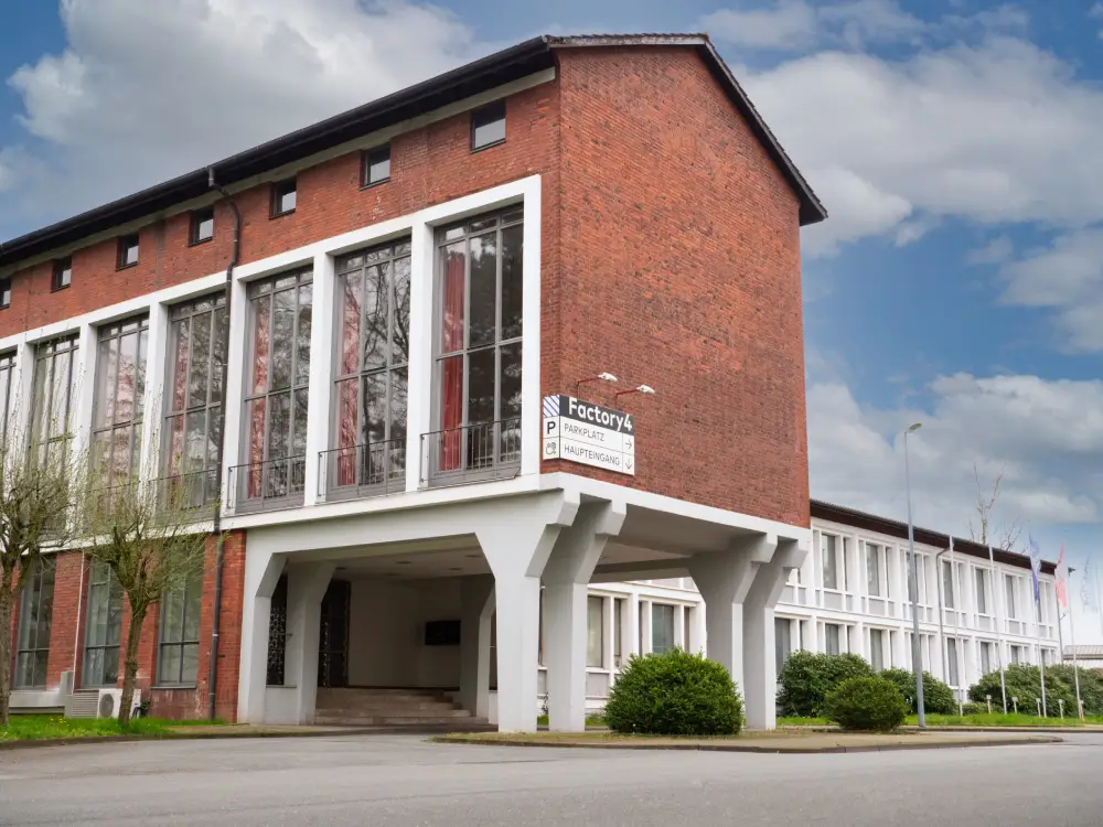 wewede-solutions_schwerte_dortmund_webdesign-webhosting_kontakt_anfahrt_gebaeude-factory4 Das Firmengebäude von GrabGoose: ein modernes Backsteingebäude mit großen, weißen Fensterrahmen. An der Ecke ist ein Schild mit der Aufschrift „Factory4“ sowie Hinweisen zum Parkplatz und Haupteingang angebracht. Der Himmel ist leicht bewölkt.